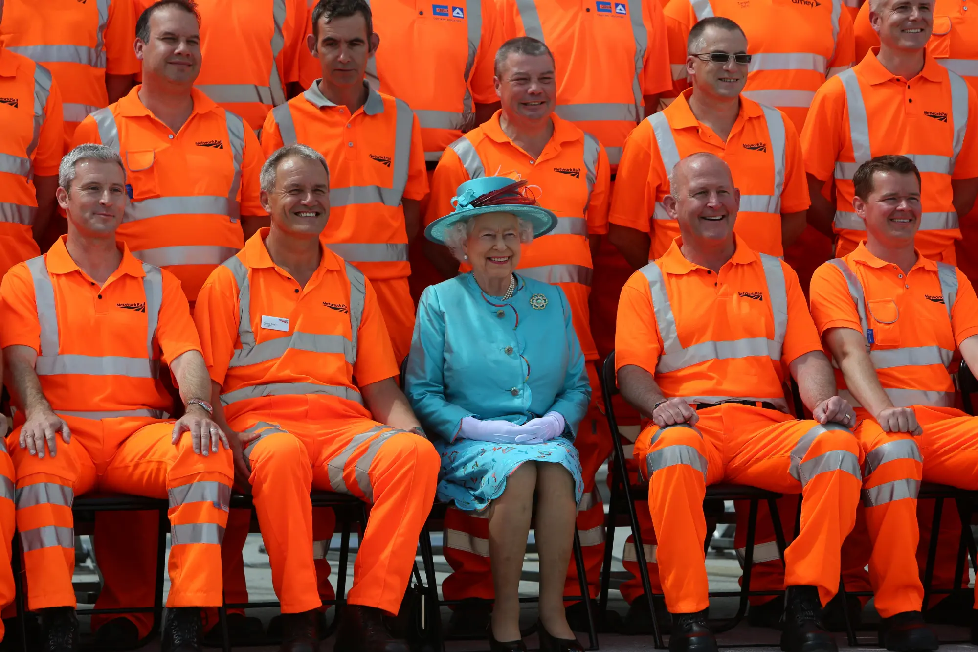 Group photo of construction workers and managers in orange rail safety jumpsuits. Queen Elizabeth is seated with the workers.