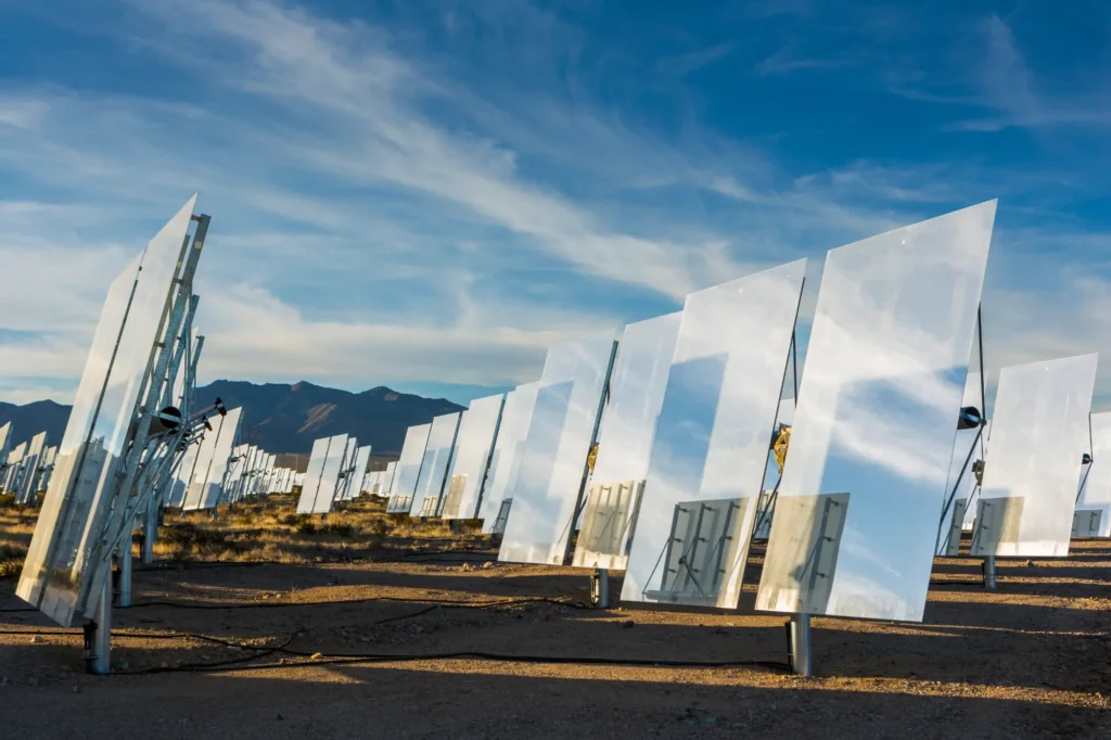 Rows of (heliostats, mirror, mirrors, solar panel, solar field) line the desert floor. They reflect the sunlight and blue sky. Ivanpah Solar Project, desert terrain, solar, energy, power